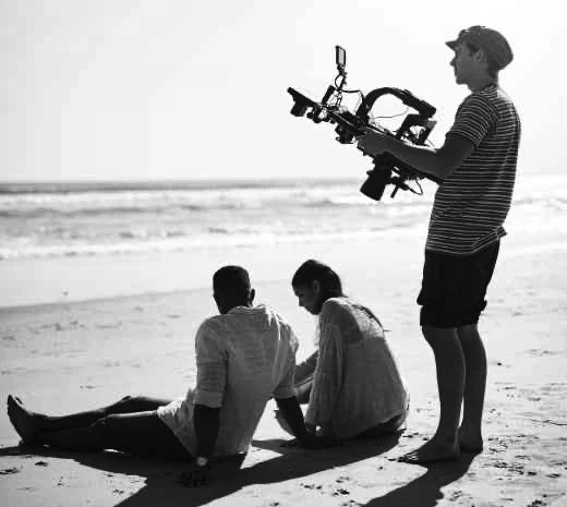 A camera operator films performers on a beach.